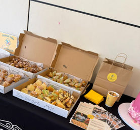 Indoor tasting table with open boxes of assorted pastries (bombolone, croissants and cream-filled treats), yellow napkins, flyers and a brown paper bag on a black tablecloth beside a pink frosted cake.
