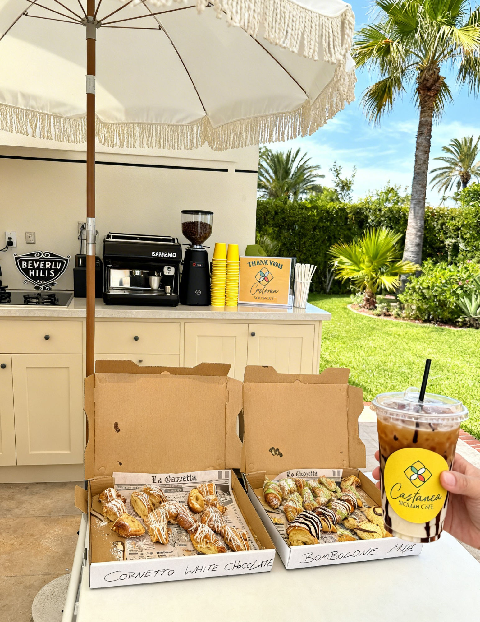 Sunny palm-lined outdoor patio coffee station with two open boxes of Italian pastries (cornetti and bomboloni) drizzled with chocolate and a hand holding an iced coffee.