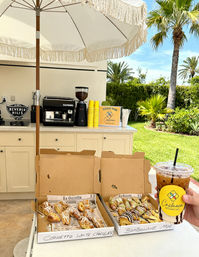 Sunny palm-lined outdoor patio coffee station with two open boxes of Italian pastries (cornetti and bomboloni) drizzled with chocolate and a hand holding an iced coffee.