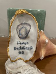 Beachy oyster shell with gold-edged rim, blue pearl illustration and custom "Bachelorette 2026" lettering — fun, personalized party keepsake displayed on a conch shell stand.
