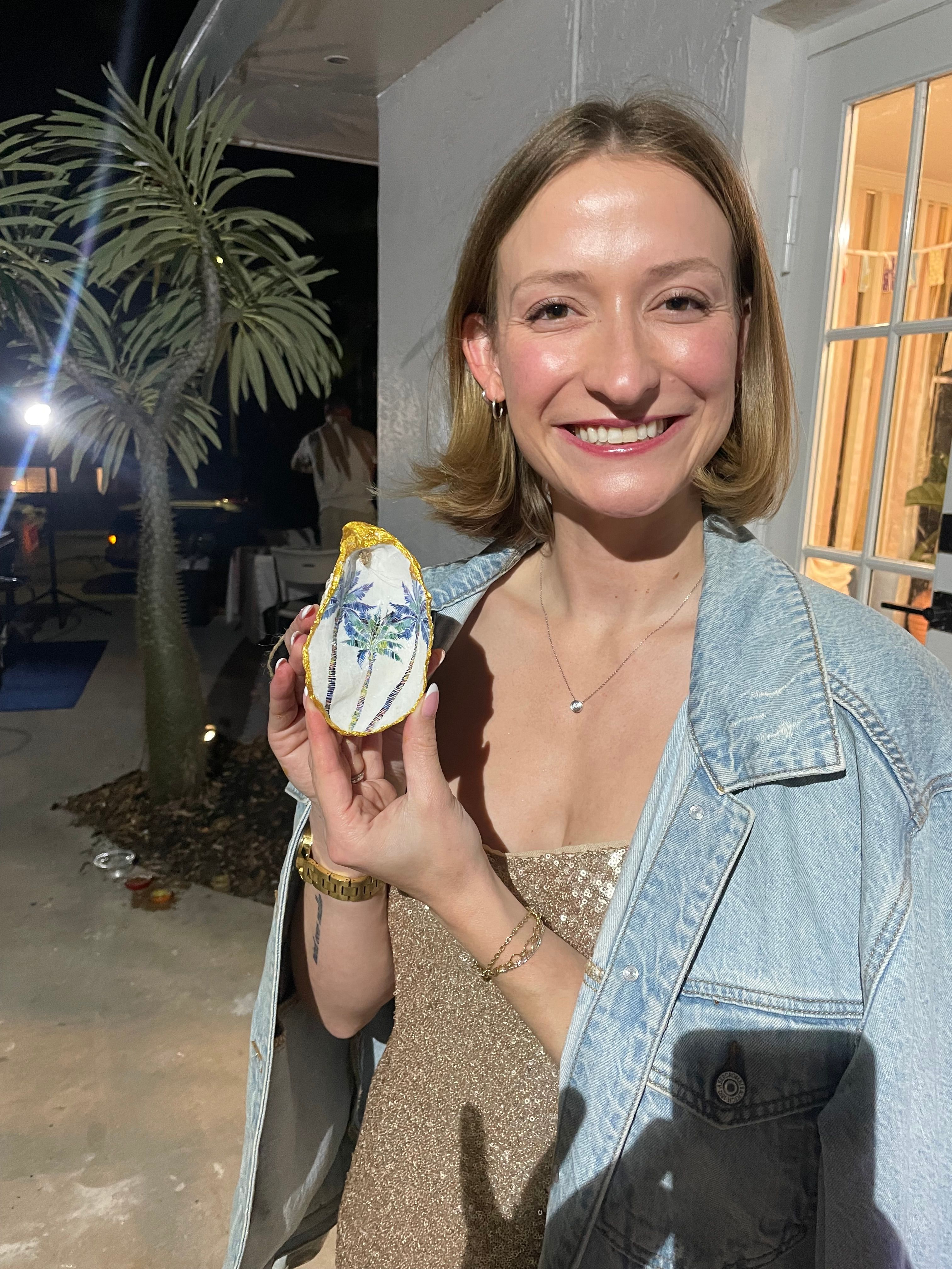 Smiling woman on an outdoor patio at night holding a hand-painted oyster shell with blue palm trees; wearing a denim jacket over a sparkly dress, tropical palm tree visible in background.