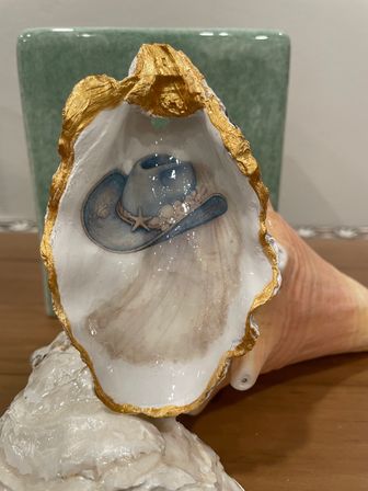 Glossy decorative seashell with gold-leaf rim featuring a painted blue cowboy hat and starfish accent, displayed on a ceramic base on a wooden table — coastal nautical home decor