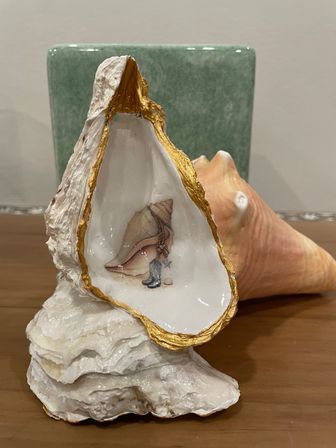 Decorative oyster shell with gilded rim and glossy painted conch motif inside, displayed on a wooden table with a pink conch shell and green tile backdrop.