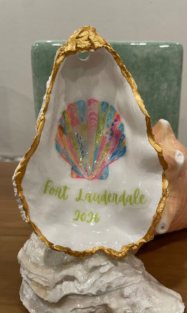 Decorative oyster shell with gold-gilded edge featuring a colorful scallop painting and text "Fort Lauderdale 2026," displayed on a white shell stand against a green tile backdrop.