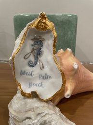 Gold-rimmed oyster shell souvenir with a painted seahorse in a cowboy hat and lasso, reading 'West Palm Beach', displayed on a seashell base with a conch shell backdrop.