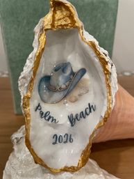 Decorative oyster shell souvenir with gold-rimmed edge, painted interior showing a blue cowboy hat, small seashells and script reading "Palm Beach 2026" on display