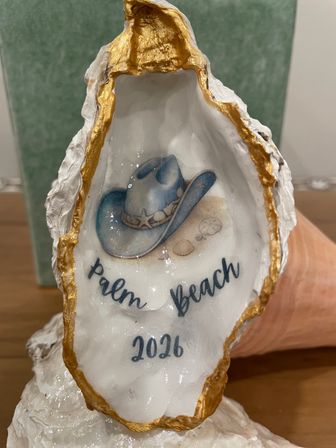 Decorative oyster shell souvenir with gold-rimmed edge, painted interior showing a blue cowboy hat, small seashells and script reading "Palm Beach 2026" on display