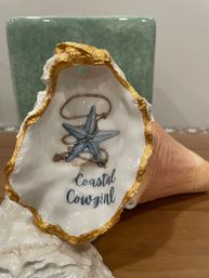 Playful decorative oyster shell with gold-gilded edge, painted interior featuring a blue starfish, coiled rope illustration and the words 'Coastal Cowgirl' — nautical coastal home decor on a wooden surface.