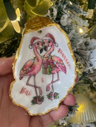 Close-up handheld gold-edged oyster shell Christmas ornament featuring two pink flamingos in Santa hats holding a wrapped gift, with festive lights and flocked tree branches in the background.