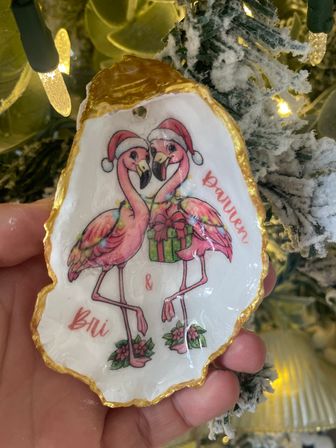 Close-up handheld gold-edged oyster shell Christmas ornament featuring two pink flamingos in Santa hats holding a wrapped gift, with festive lights and flocked tree branches in the background.