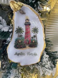 Oyster-shell Christmas ornament with gold trim featuring a red lighthouse wrapped in lights, palm trees and “Jupiter, Florida” text, hanging on a snowy, ribboned holiday tree.