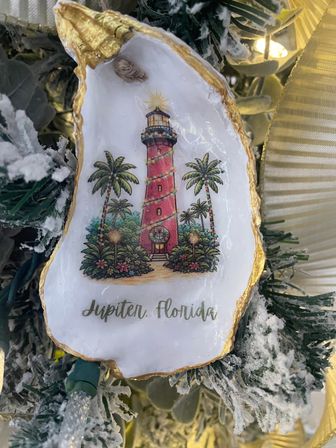 Oyster-shell Christmas ornament with gold trim featuring a red lighthouse wrapped in lights, palm trees and “Jupiter, Florida” text, hanging on a snowy, ribboned holiday tree.