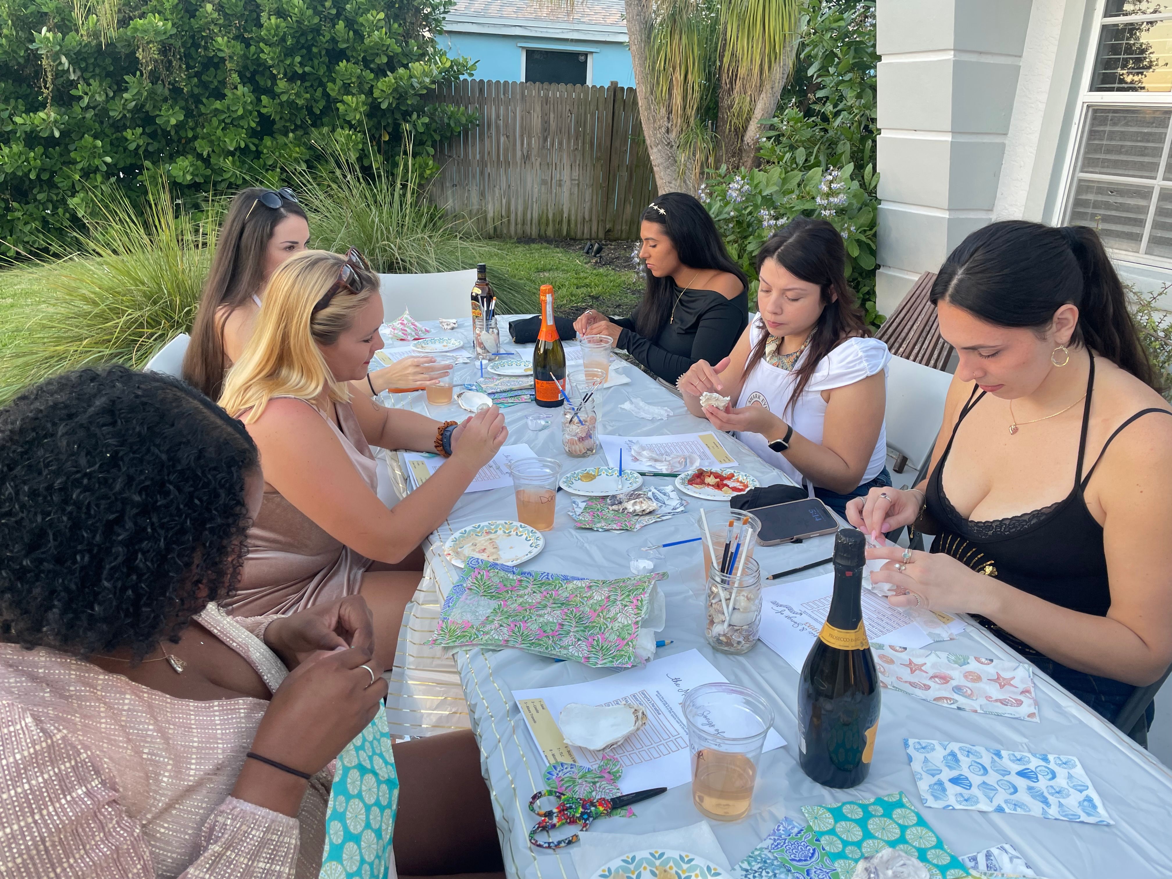 Group of women seated around a backyard garden table crafting shell art and enjoying drinks, with patterned napkins, paintbrushes and champagne bottles.
