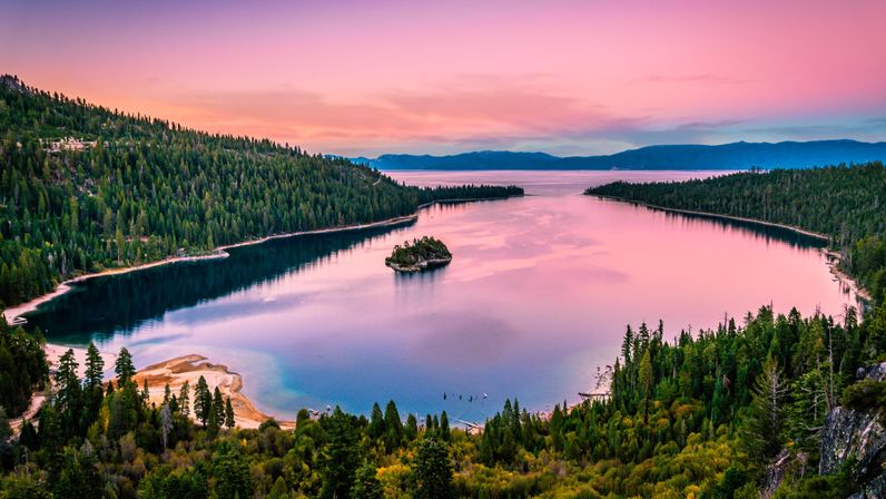 Aerial panorama of a mountain lake at pink sunset, a tiny tree‑topped island in glassy water framed by pine‑covered shores and sandy coves.