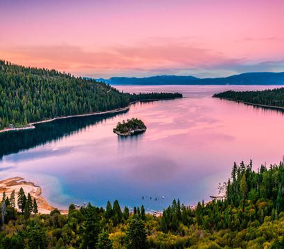 Aerial panorama of a mountain lake at pink sunset, a tiny tree‑topped island in glassy water framed by pine‑covered shores and sandy coves.