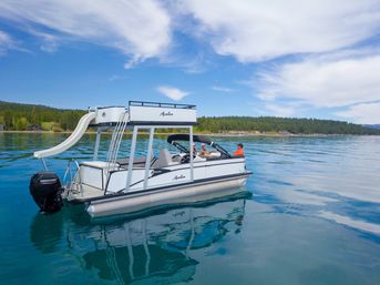 White two-deck pontoon boat with waterslide floating on a clear blue mountain lake near a pine-forested shoreline under a bright sky, three people relaxing aboard.