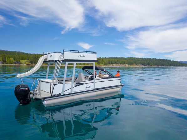 White two-deck pontoon boat with waterslide floating on a clear blue mountain lake near a pine-forested shoreline under a bright sky, three people relaxing aboard.