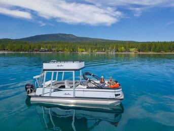 Three people relaxing on a white pontoon boat on a crystal-clear mountain lake, turquoise water reflecting a blue sky, with a pine-forested shoreline and rolling hills in the background.