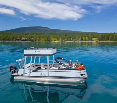 Three people relaxing on a white pontoon boat on a crystal-clear mountain lake, turquoise water reflecting a blue sky, with a pine-forested shoreline and rolling hills in the background.