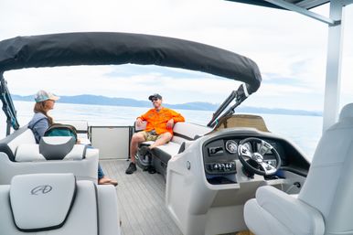 Two passengers lounging on a pontoon boat with steering console and black bimini top on a calm mountain lake with distant snow-capped peaks.