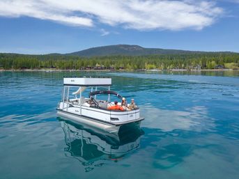 Pontoon boat with three people relaxing on a calm, clear-blue alpine lake, reflecting the sky, with a forested shoreline, cabins and distant mountains under a sunny sky.