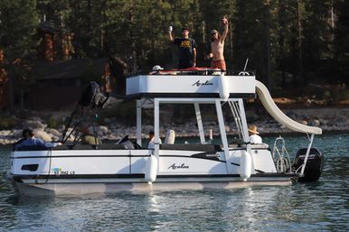 Pontoon boat with upper deck and waterslide on a calm mountain lake, people waving and relaxing near a forested rocky shoreline