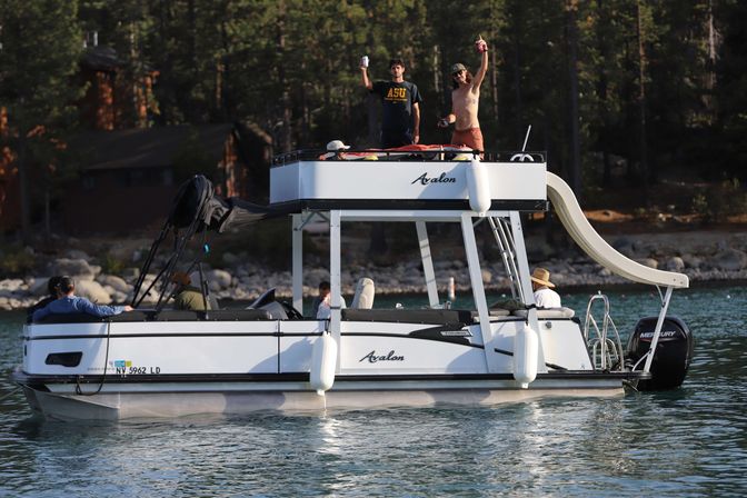 Pontoon boat with upper deck and waterslide on a calm mountain lake, people waving and relaxing near a forested rocky shoreline