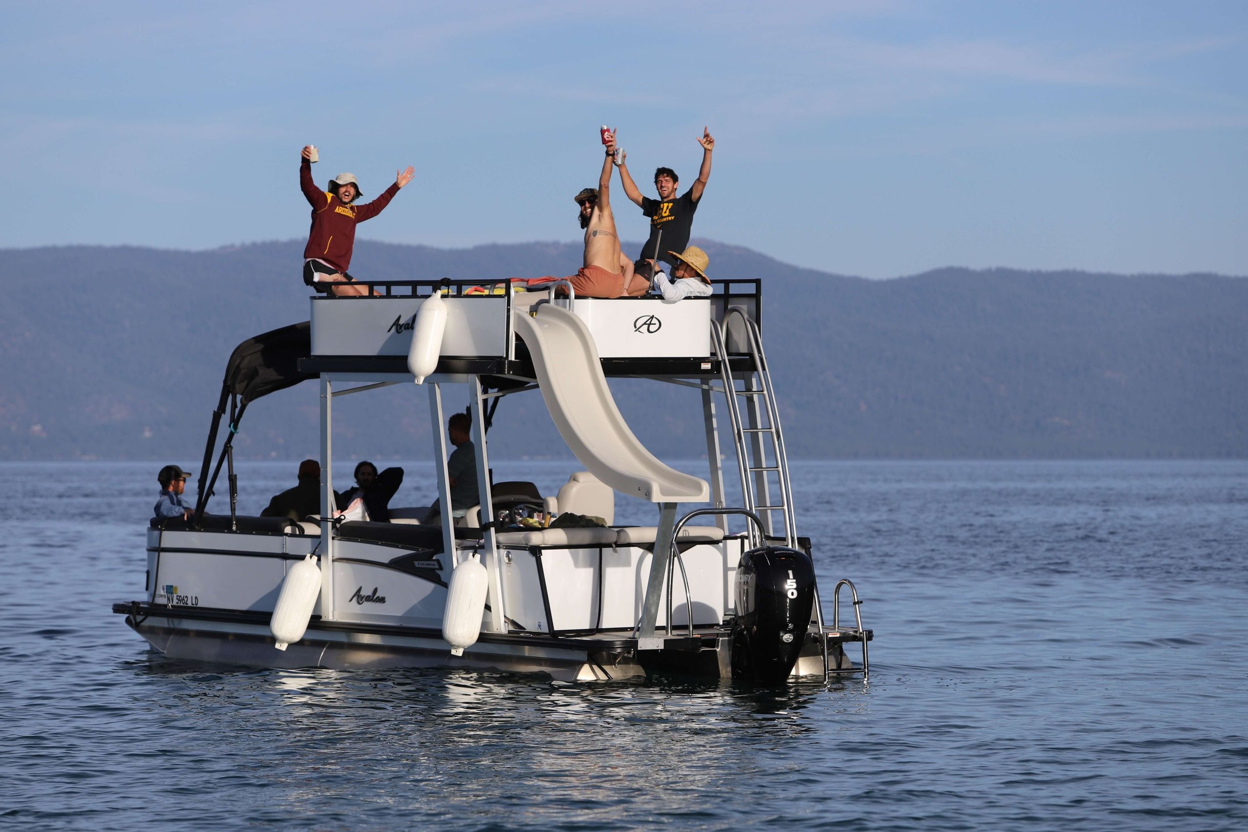 Friends cheering on a two-level pontoon boat with a rooftop slide, floating on a calm mountain lake under a clear blue sky