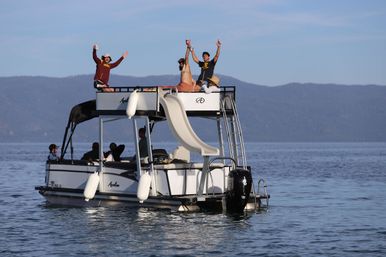 Friends cheering on a two-level pontoon boat with a rooftop slide, floating on a calm mountain lake under a clear blue sky