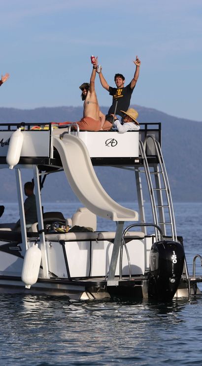 Friends cheering on a two-level pontoon boat with a rooftop slide, floating on a calm mountain lake under a clear blue sky