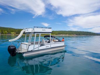 White pontoon boat with slide and outboard motor floating on a clear, calm mountain lake near a pine-lined shoreline under a blue sky with wispy clouds.