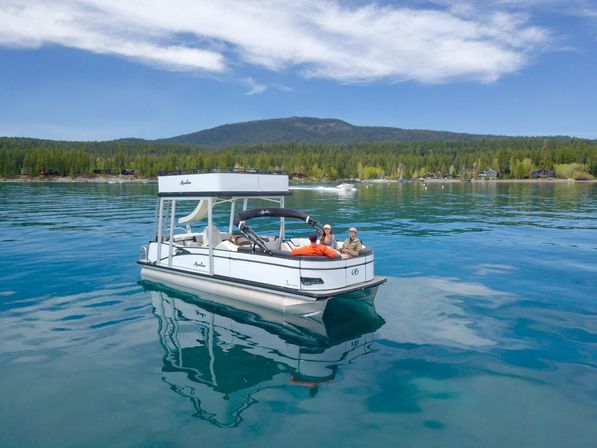 White pontoon boat with people relaxing on a crystal-clear mountain lake, turquoise water reflecting a forested shoreline and blue sky with wispy clouds.