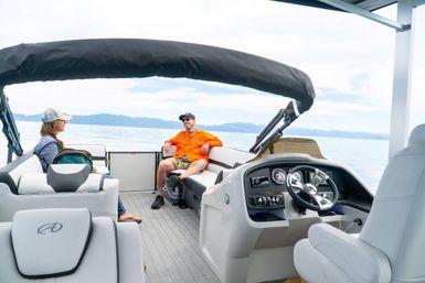 Two adults relaxing on a pontoon boat under a black canopy, modern helm and steering wheel visible, calm alpine lake and distant mountains, chill boating vibe.