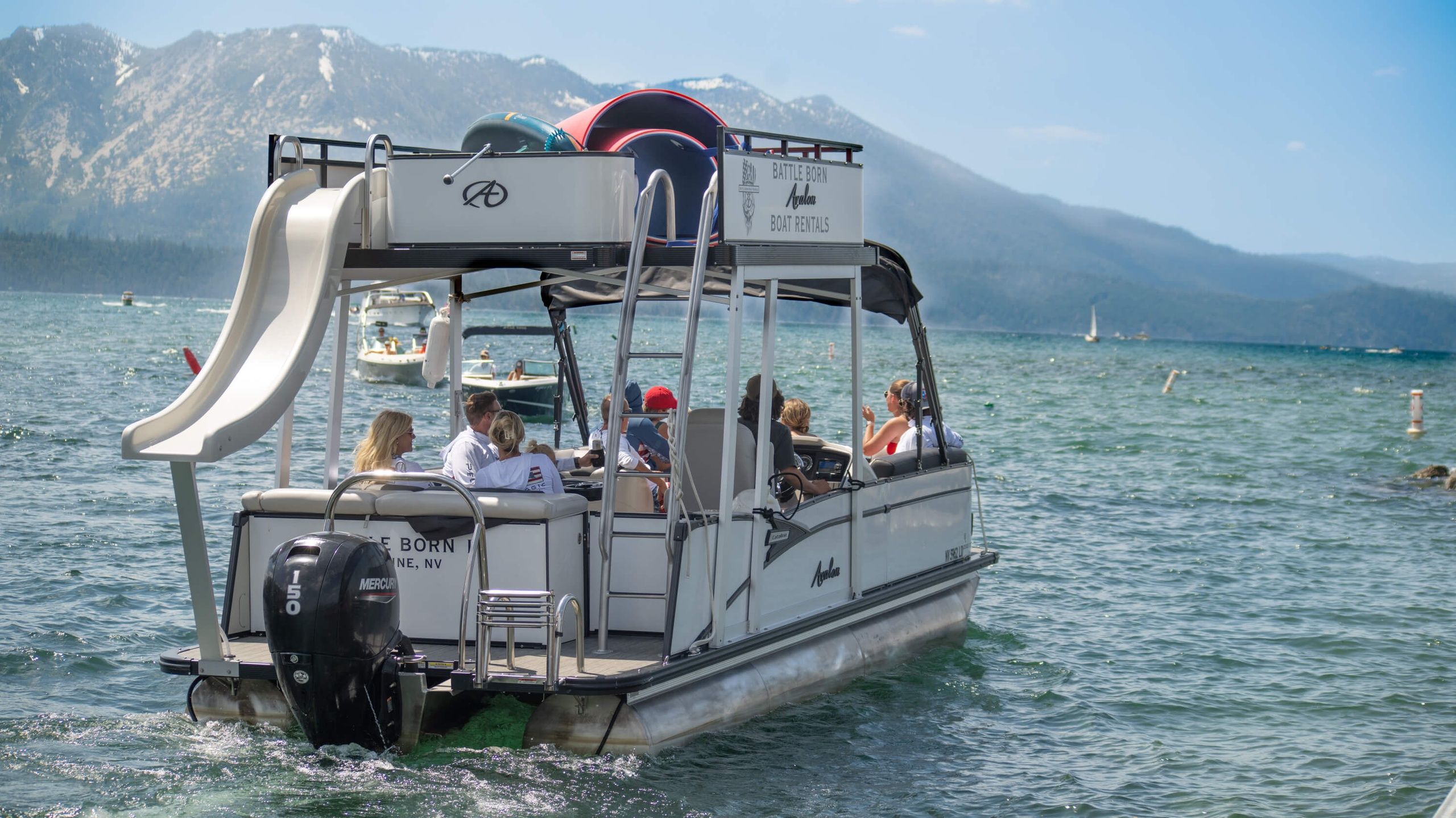 Pontoon boat with rooftop slide and outboard motor carrying a group of people on a sunlit alpine lake, forested mountains rising in the background — summer boating and waterside fun.