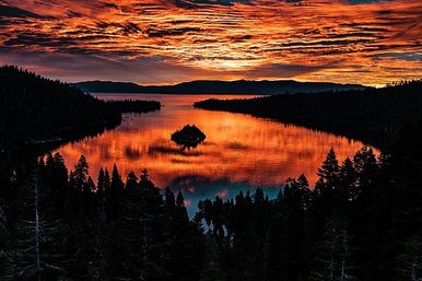 Fiery orange-red sunset over an alpine mountain lake with a small island centered in glassy water, silhouetted pine forests and dramatic cloud reflections.