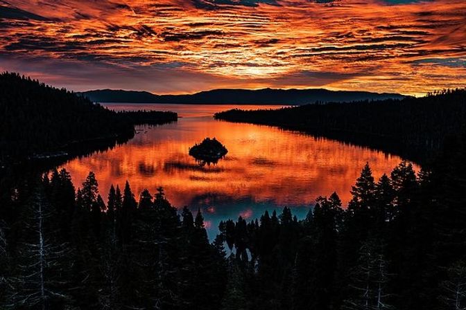 Fiery orange-red sunset over an alpine mountain lake with a small island centered in glassy water, silhouetted pine forests and dramatic cloud reflections.