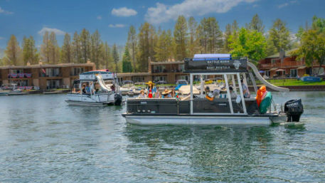 Pontoon rental boats with a waterslide and colorful kayaks carrying groups of people across a pine‑lined lake in front of waterfront vacation condos on a sunny summer day.