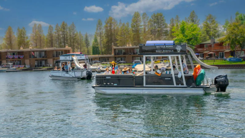 Pontoon rental boats with a waterslide and colorful kayaks carrying groups of people across a pine‑lined lake in front of waterfront vacation condos on a sunny summer day.