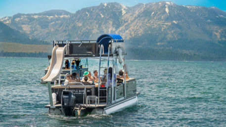 Pontoon party boat with rooftop water slide and a group of people enjoying a sunny day on a clear blue mountain lake with forested shoreline and rugged peaks in the background.