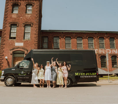 Cheerful group of women in summer dresses posing and waving in front of a black party van parked by a historic red-brick industrial building on a sunny urban street