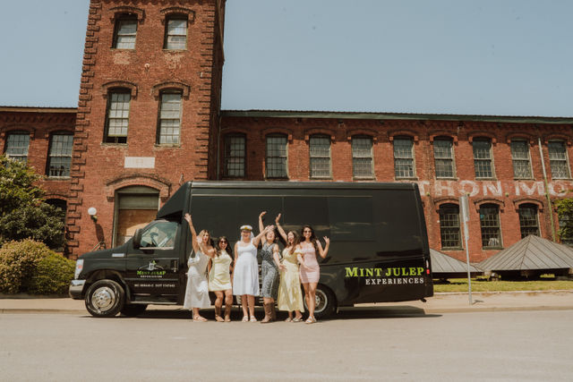 Cheerful group of women in summer dresses posing and waving in front of a black party van parked by a historic red-brick industrial building on a sunny urban street