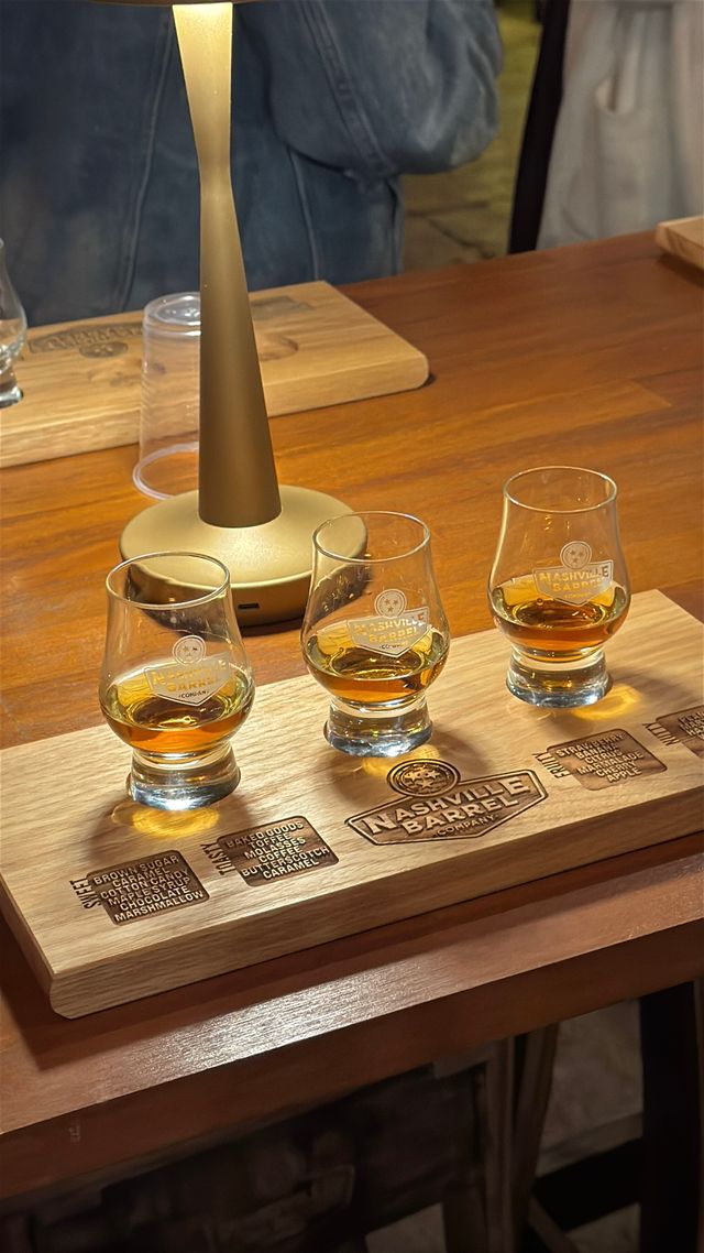 Three-glass whiskey flight with amber pours on an engraved wooden tasting board under a warm table lamp on a wooden bar, cozy tasting scene.