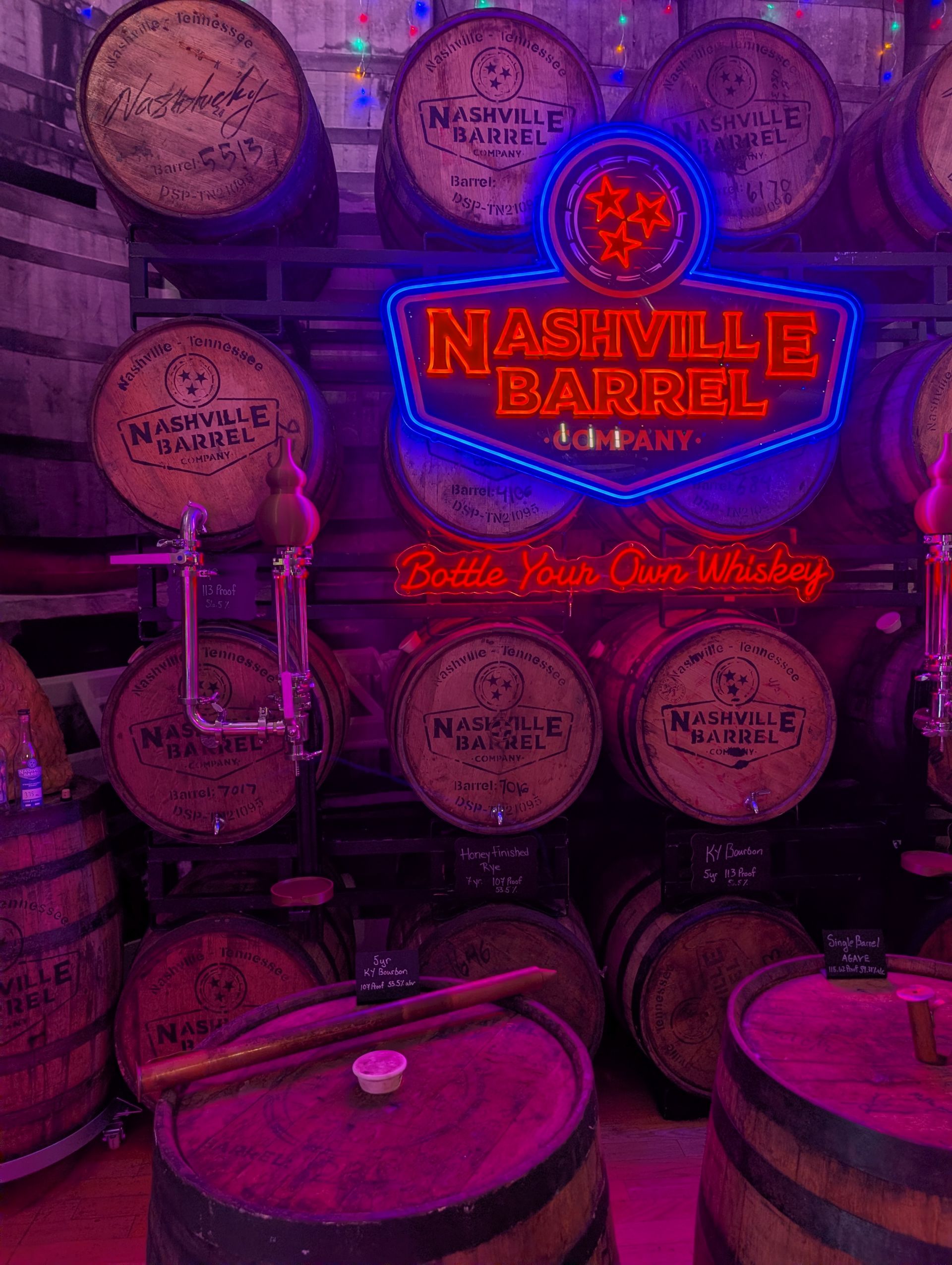 Neon-lit Nashville whiskey barrel display with stacked oak barrels, vintage taps and scoops, and a bright blue-and-red neon sign promoting bottle-your-own whiskey.