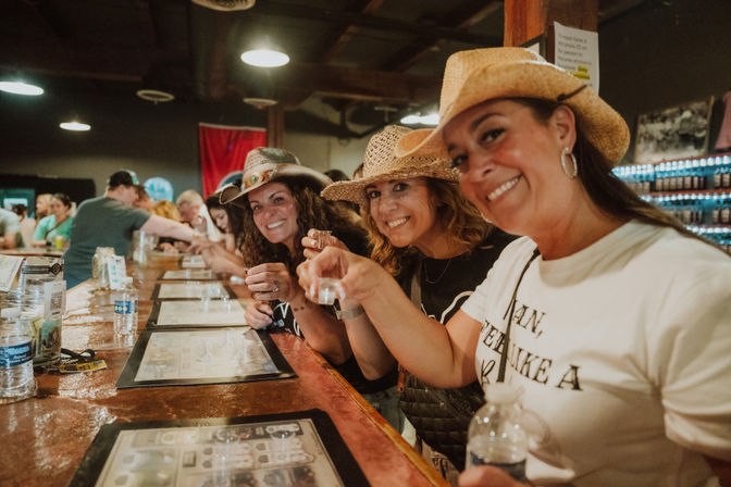 Three smiling women in straw cowboy hats clinking small tasting glasses at a lively indoor bar or tasting room, wooden counter and menus in the foreground and a crowd in the background.