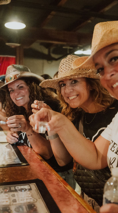 Three smiling women in straw cowboy hats clinking small tasting glasses at a lively indoor bar or tasting room, wooden counter and menus in the foreground and a crowd in the background.