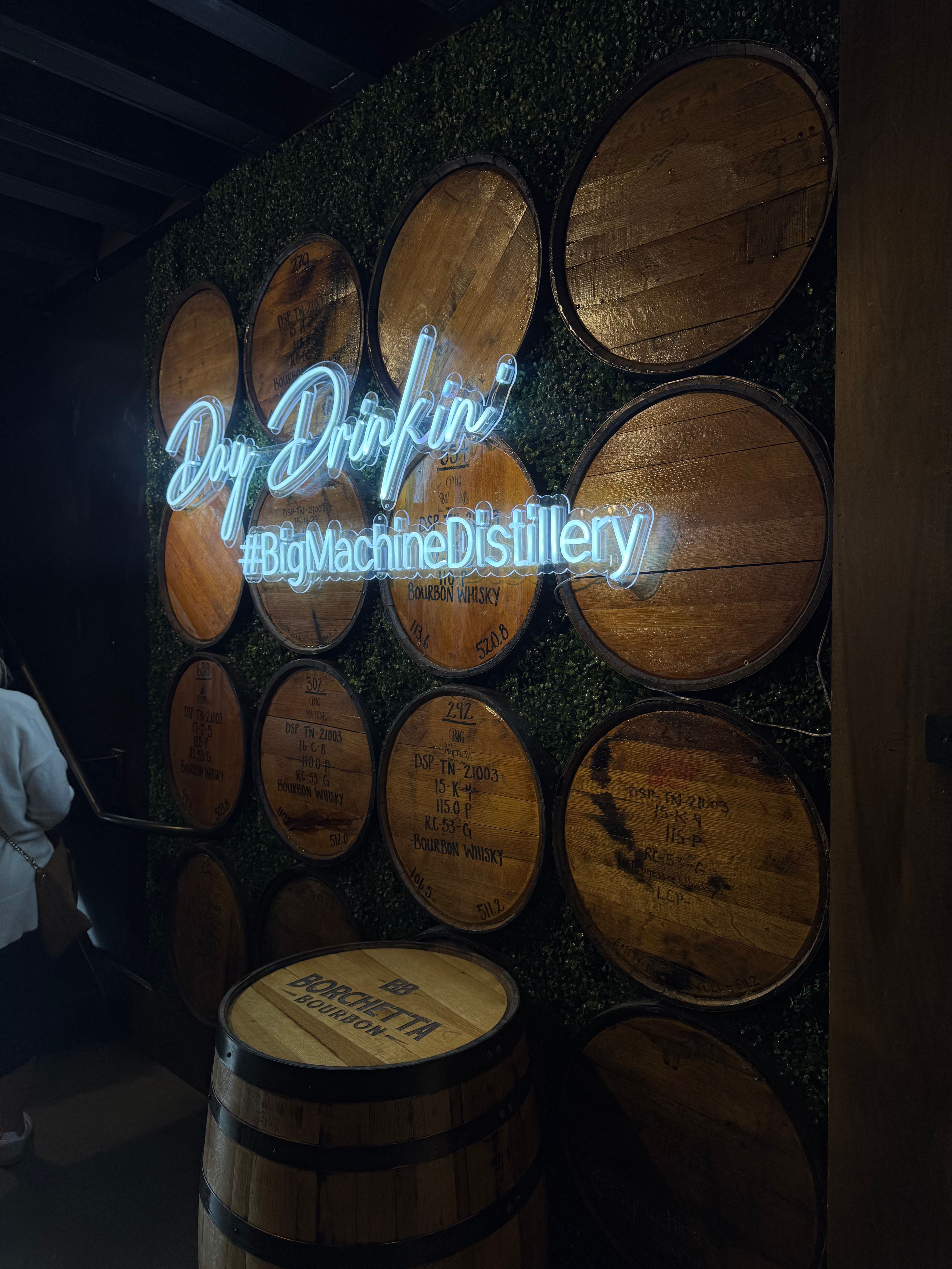 Neon blue “Day Drinkin’” sign glowing over stacked bourbon barrels and a barrel-top table in a dim indoor distillery tasting room.