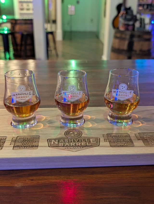Three amber whiskey samples in Glencairn-style glasses on an engraved wooden tasting flight paddle at a Nashville tasting room with colorful LED reflections