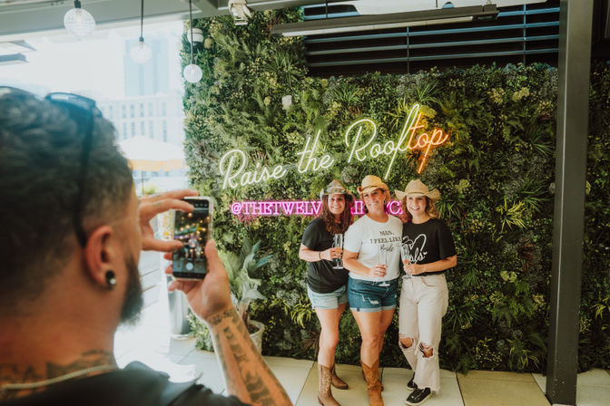 Three friends in cowboy hats holding drinks and smiling in front of a lush living wall with a neon “Raise the Rooftop” sign at an urban rooftop bar, captured on a smartphone.