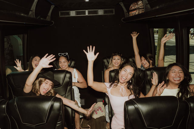 Cheerful group of friends waving and laughing inside a black-leather seat party van during a daytime city ride.