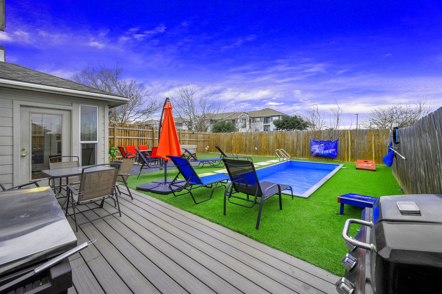 Inviting suburban backyard patio with a small rectangular plunge pool on bright artificial turf, several black lounge chairs and a vibrant orange umbrella, wooden privacy fence, deck dining table and barbecue grill under a vivid blue sky.